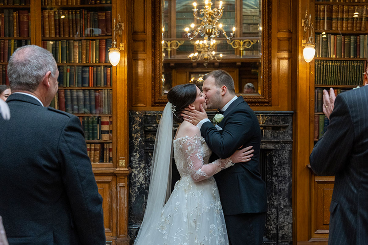 A bride and groom kiss during their ceremony in a room with wood paneling and lots of books on shelves