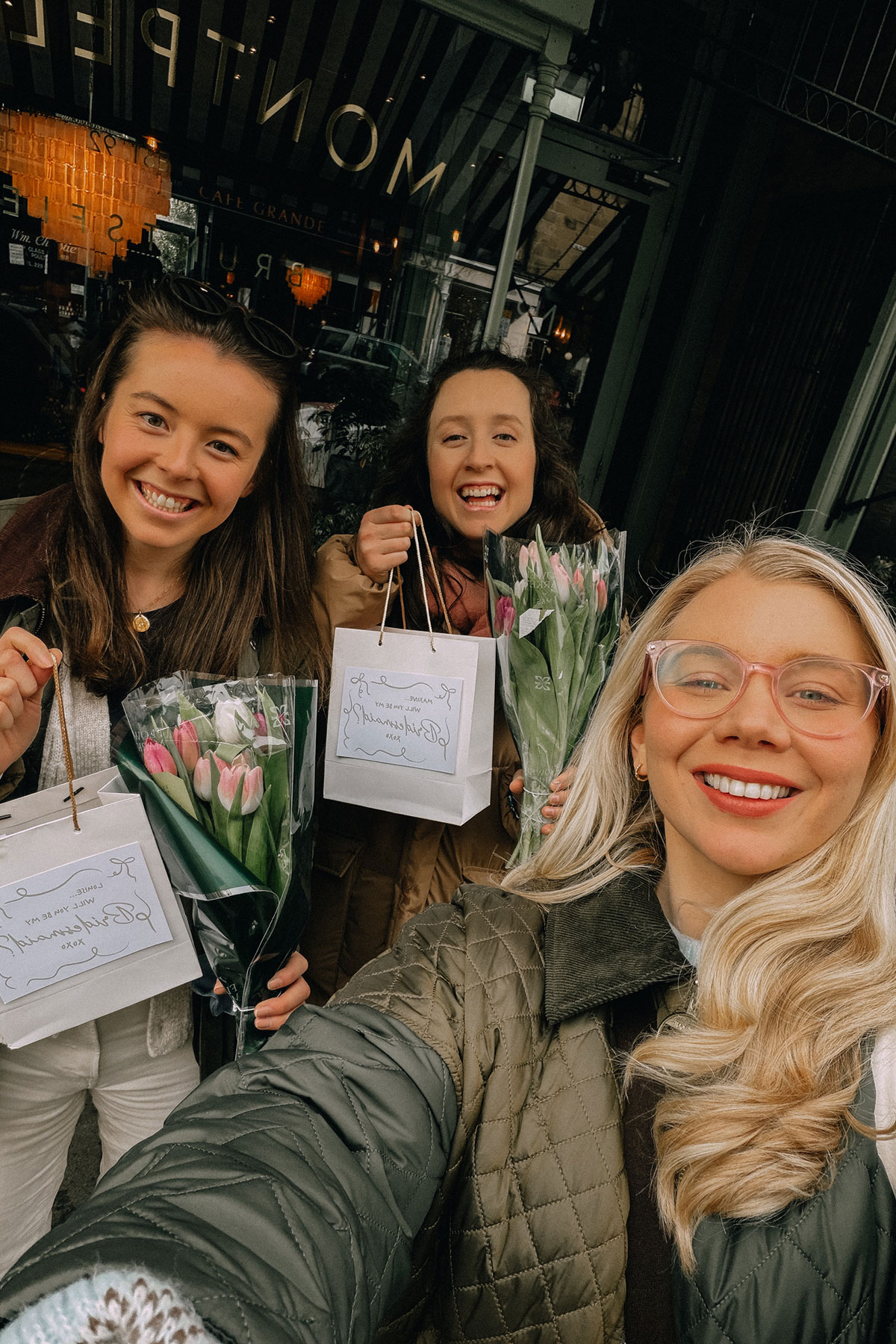 Three women smiling outside cafe holding tulips and gift bags during city outing