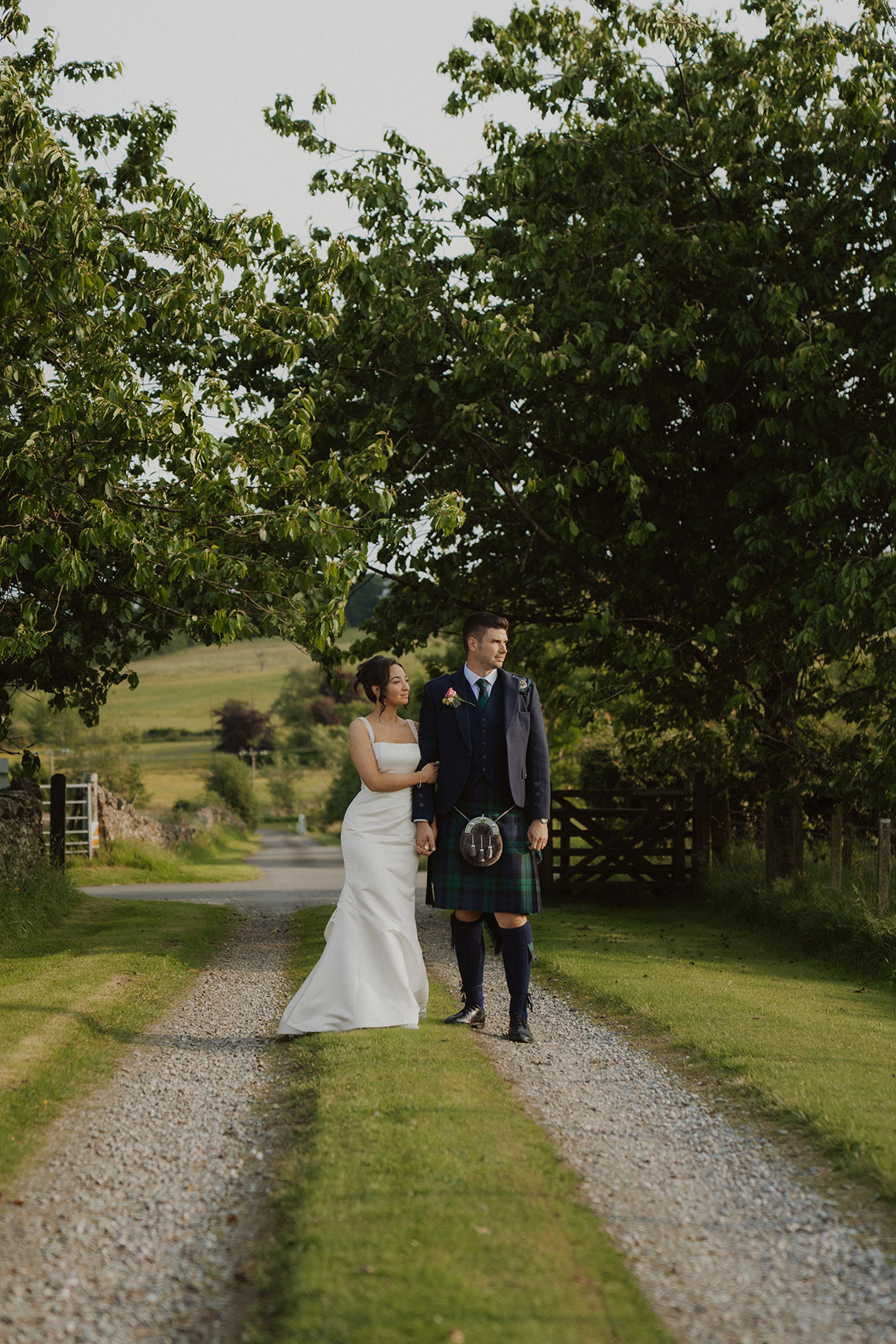 Bride and groom walking hand in hand along tree-lined gravel driveway at Scottish countryside wedding venue, groom wearing traditional tartan kilt