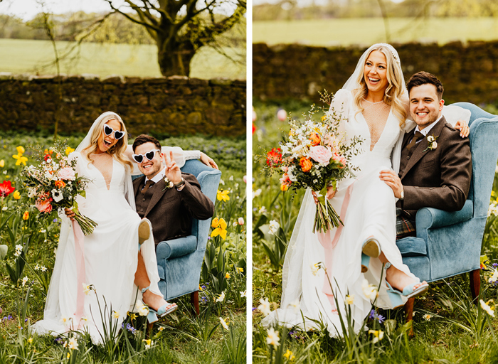 a happy bride and groom sitting on a blue velvet armchair in a flower-filled garden. The bride holds a large pink and peach pastel bouquet of flowers