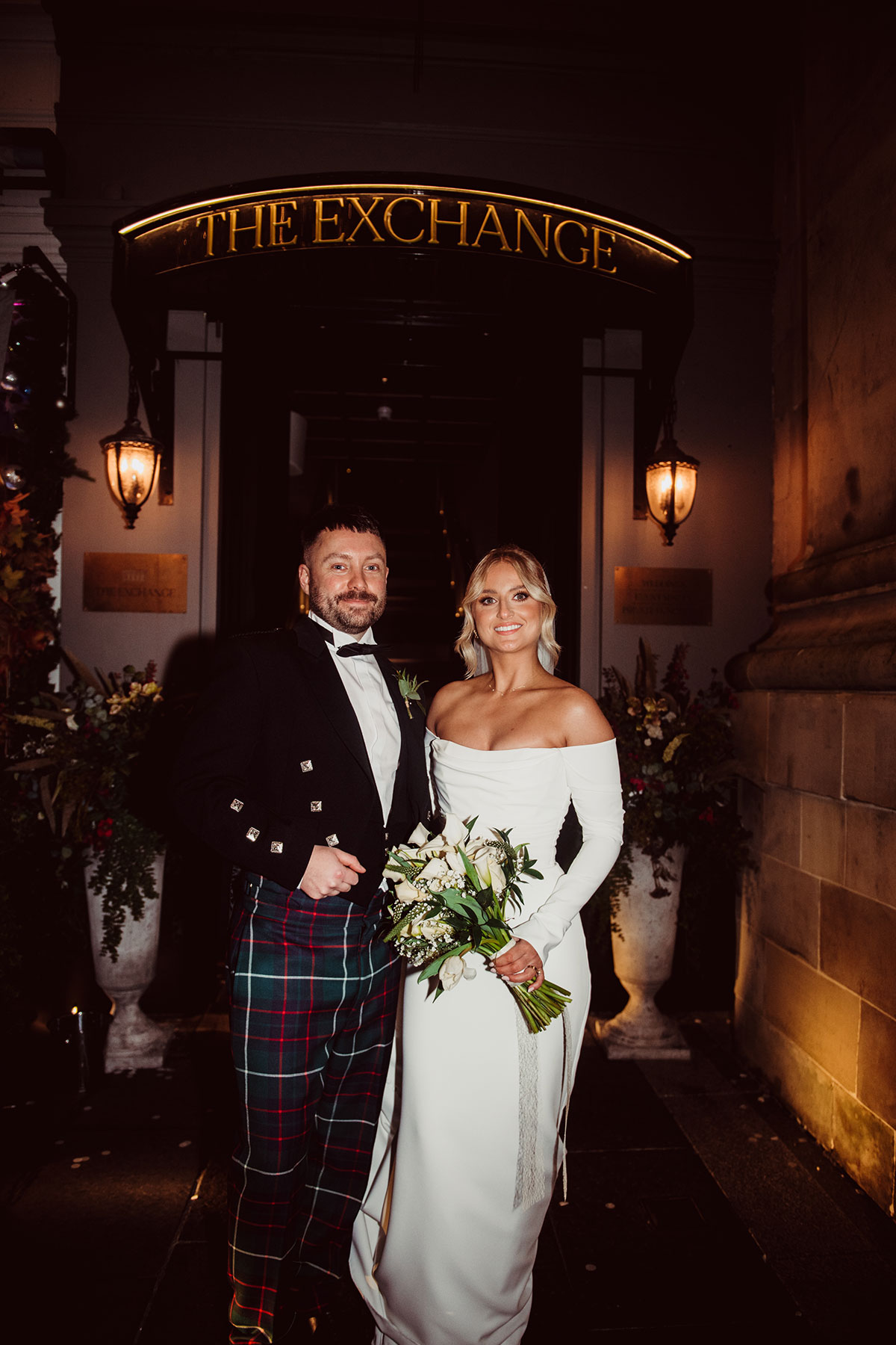 Bride and groom posing outside a venue entrance at night, standing beneath a lit sign and flanked by large floral arrangements