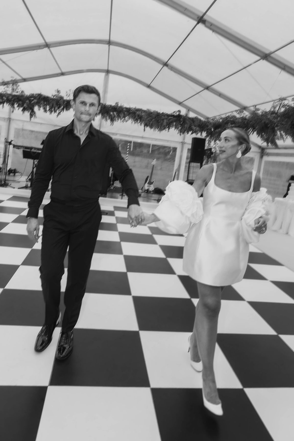 Black and white photo of newlyweds holding hands on a checkered dance floor inside a wedding marquee