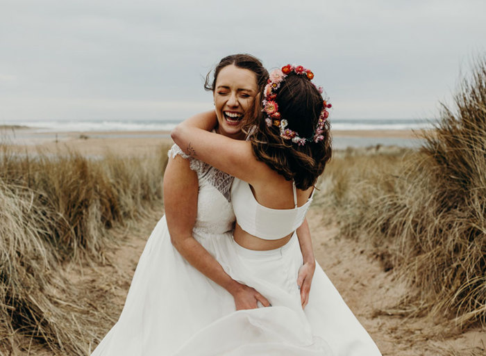 two brides hugging on a beach with sand, sea and scrubby grass in the background