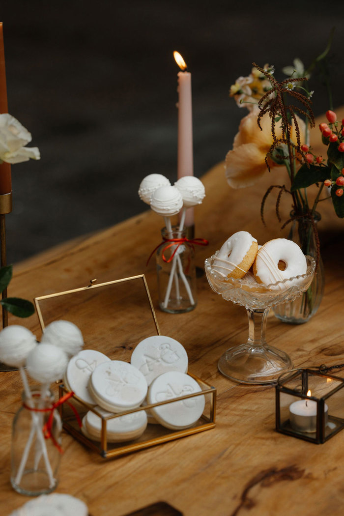 glass containers display a variety of white icing treats including cake pops, ring donuts and imprinted iced biscuits while tea lights and coloured candles sit around them on the wooden dining table