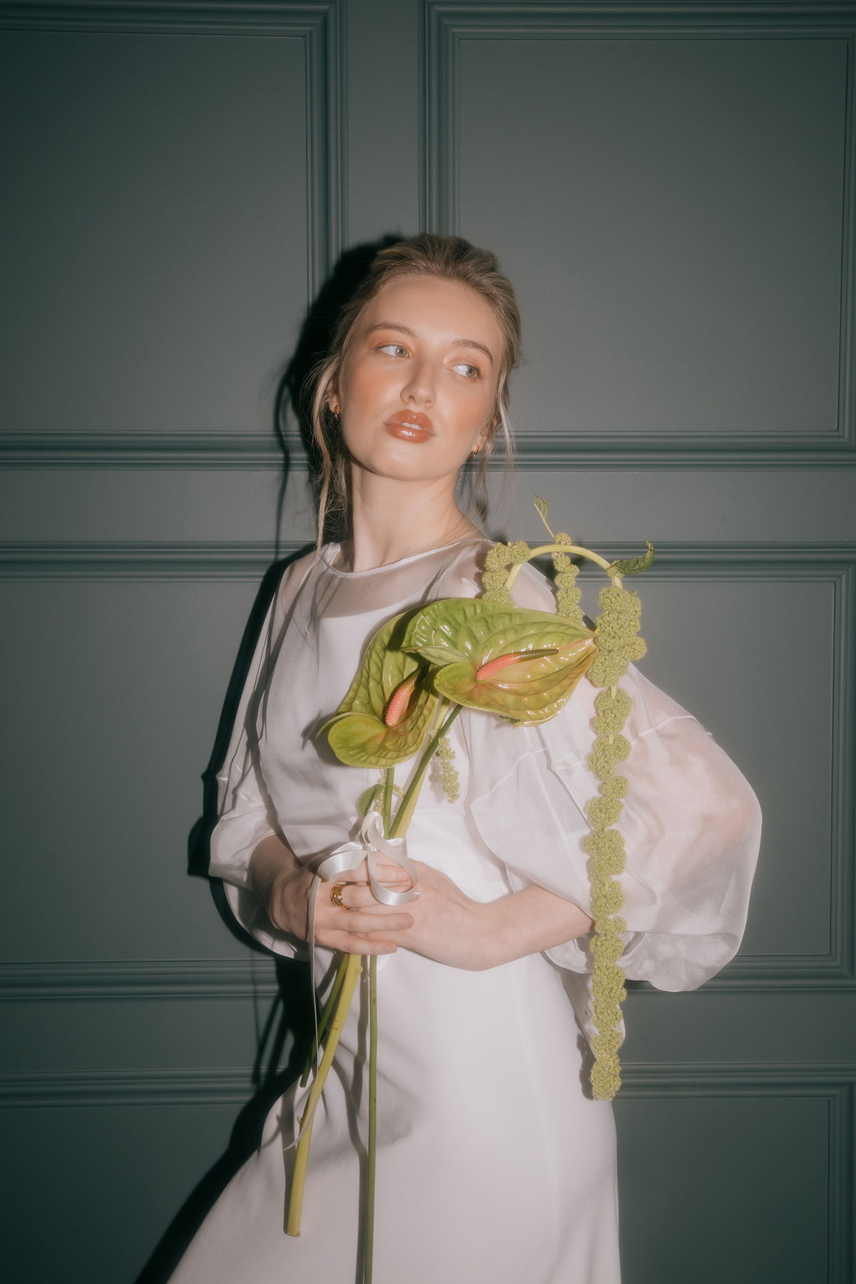 A bride in a white gown poses against a dark teal panelled wall, holding a bouquet of green anthuriums.