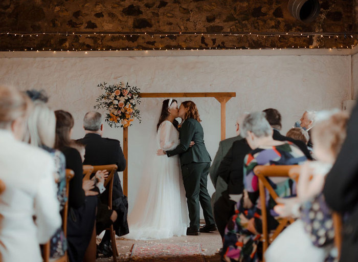 two brides kissing during a wedding ceremony at Cow Shed Crail. They stand below a wooden arch with a flower posy on the top left corner and rows of wedding guests sitting on wooden cross back chairs look on