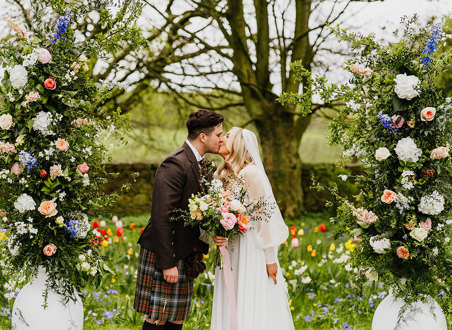 a bride and groom kissing in a garden filled with abundant colourful flowers. Two large white urns filled with tall pastel and foliage flower arrangements flank them 
