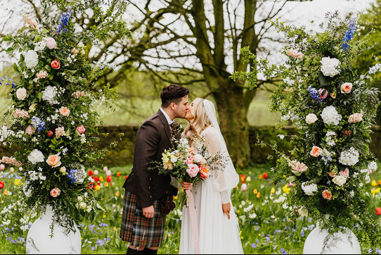 a bride and groom kissing in a garden filled with abundant colourful flowers. Two large white urns filled with tall pastel and foliage flower arrangements flank them 