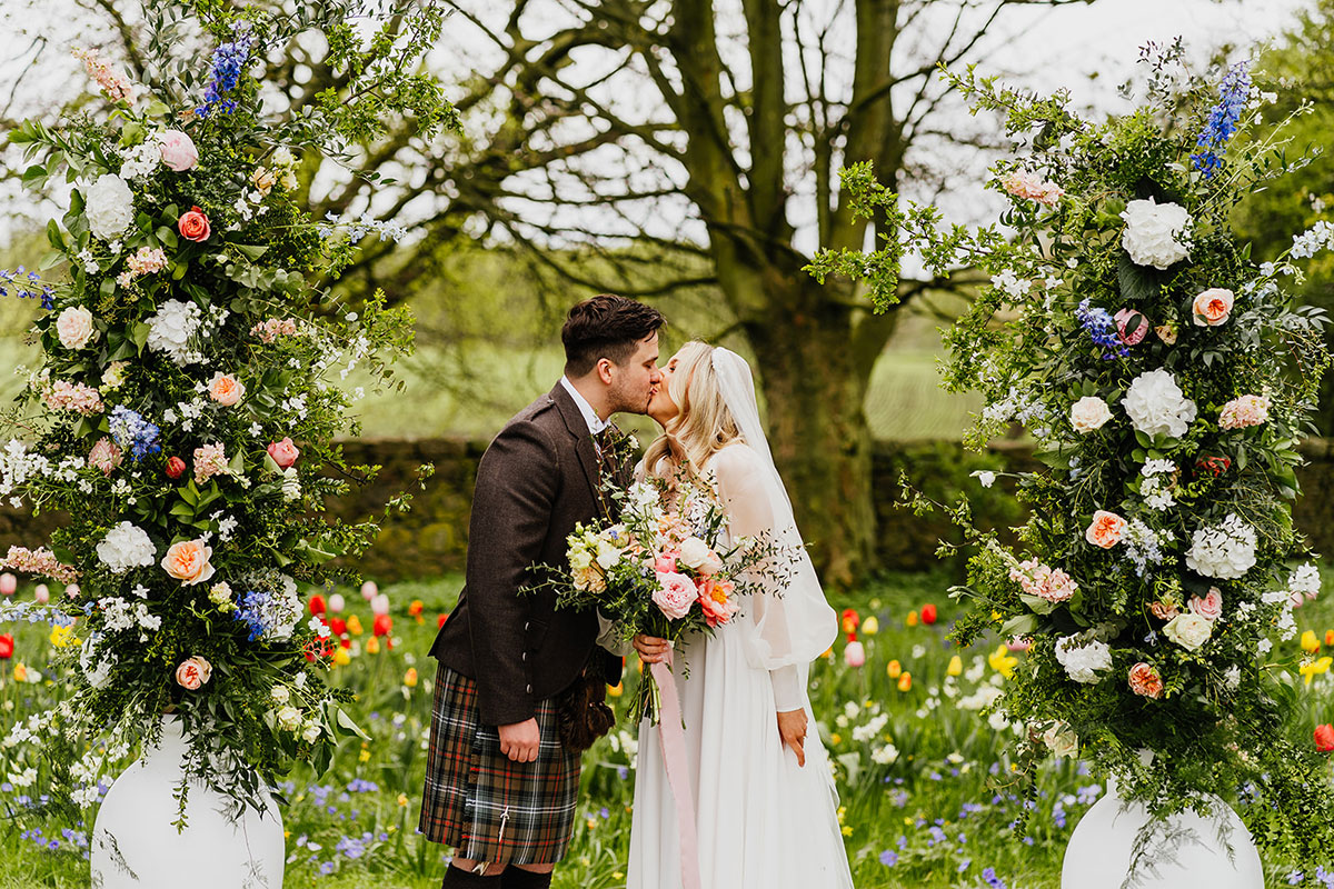 a bride and groom kissing in a garden filled with abundant colourful flowers. Two large white urns filled with tall pastel and foliage flower arrangements flank them