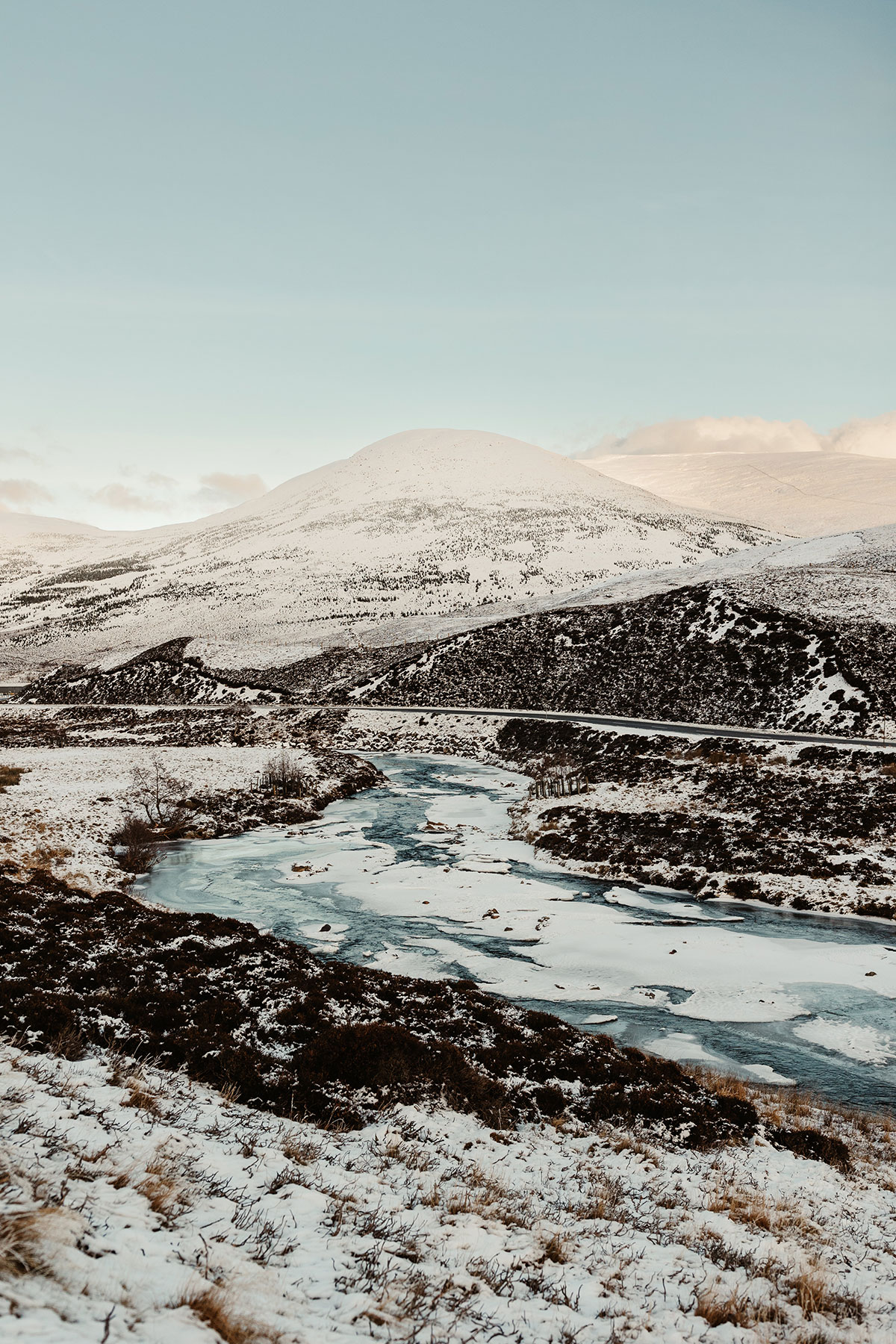 Snow-covered Highland landscape with rolling hills and a partially frozen river beneath a pale winter sky