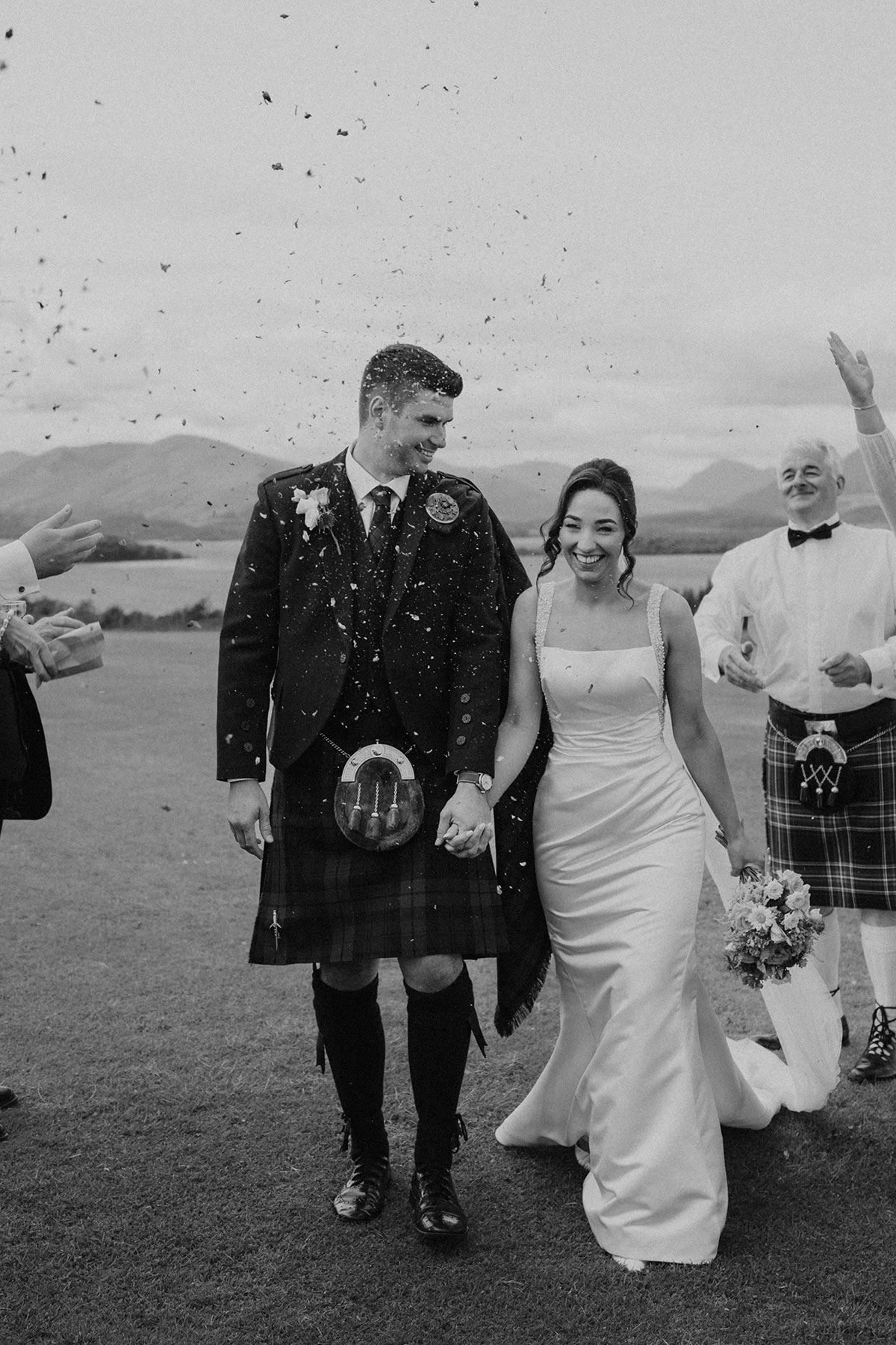 Black and white confetti moment as Scottish bride and groom walk hand in hand with loch and mountains in the background