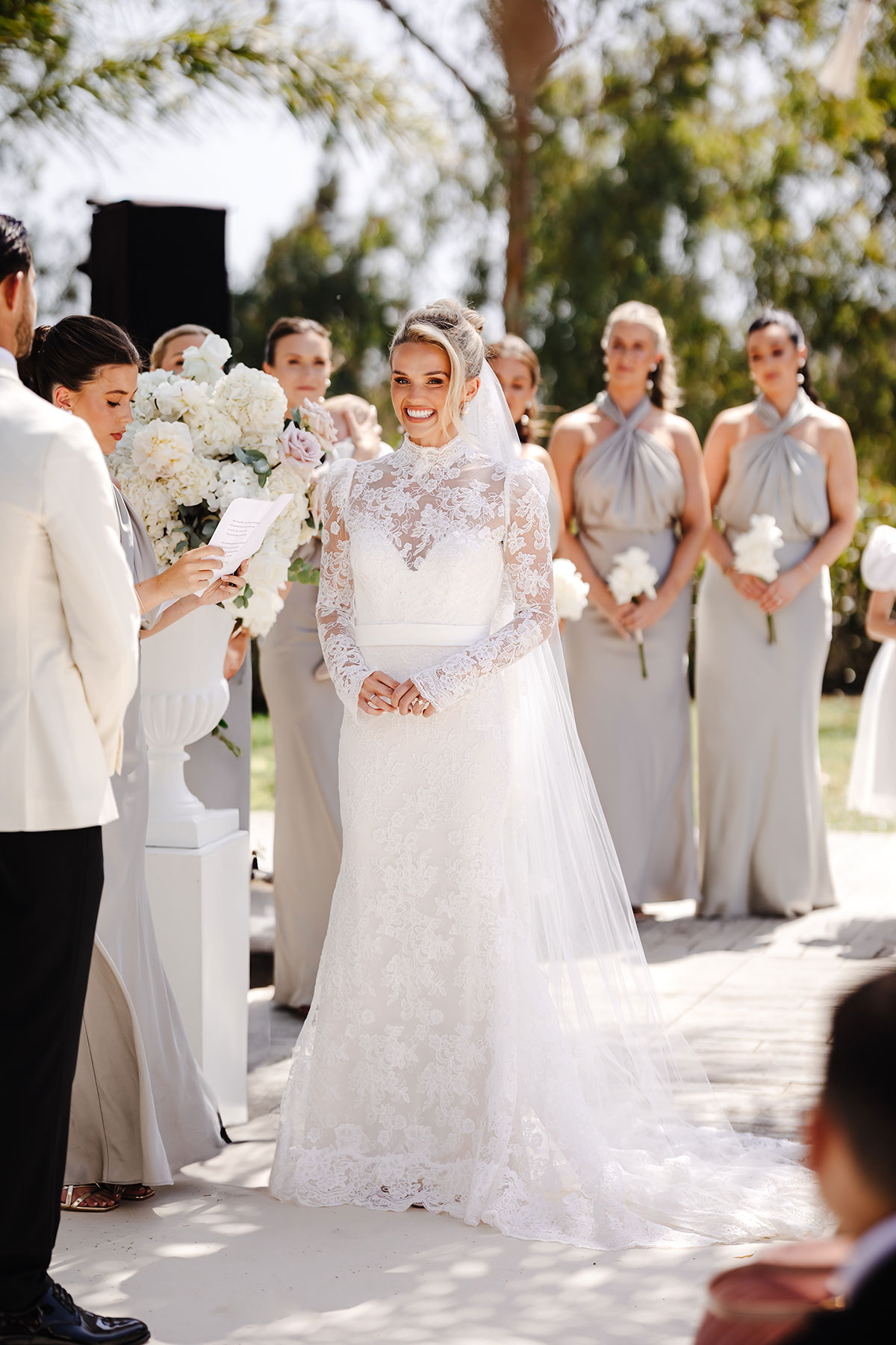 Bride smiles as a bridesmaid reads vows, with bridesmaids in silver dresses behind her