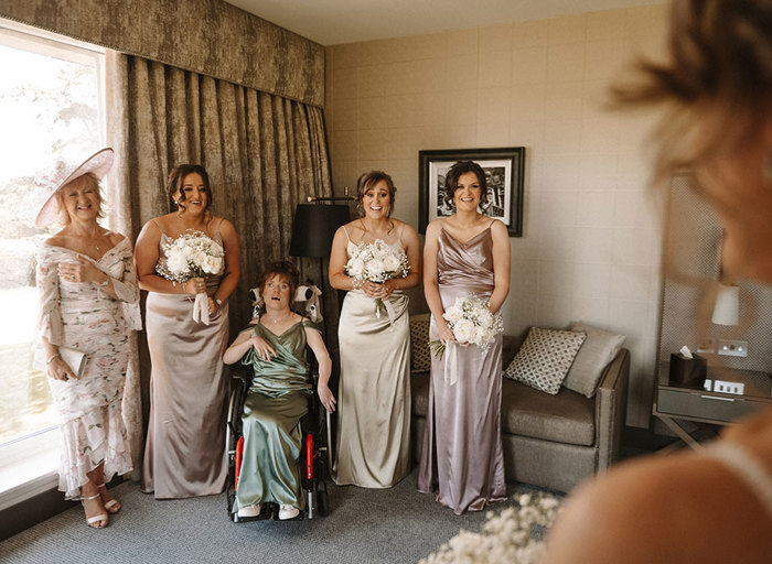 four bridesmaids wearing satin dresses and a person wearing a pale pink floral dress look at a bride wearing her wedding dress for the first time with happy and surprised expressions on their faces. One of the bridesmaids is in a wheelchair.