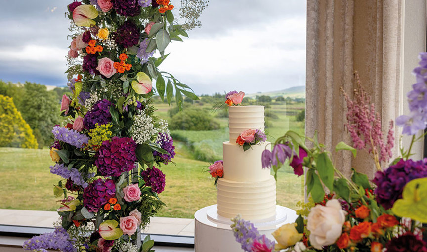 an ivory tiered buttercream cake with rainbow-coloured flowers in front of a window