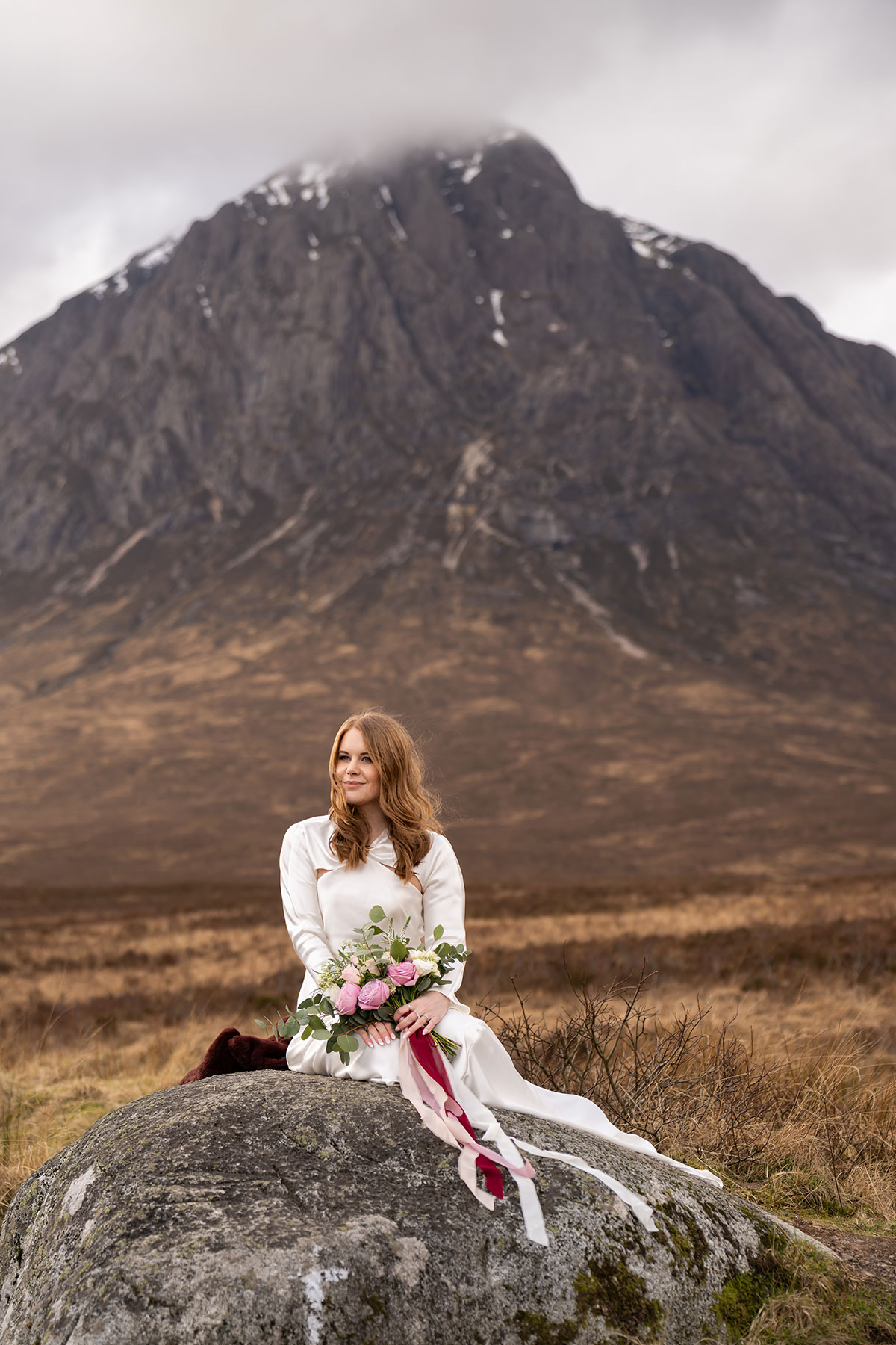 Bride sitting on a large rock with her bouquet in front of Buachaille Etive Mòr during a Glencoe elopement photoshoot