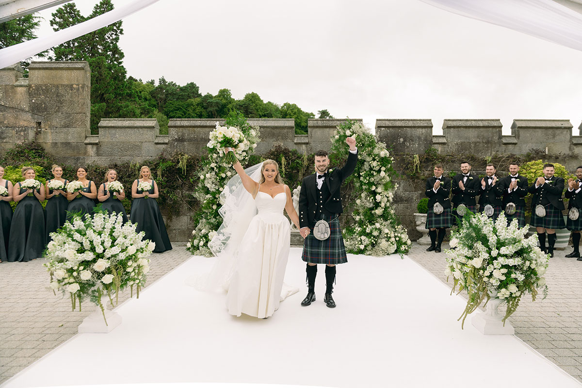 Newlyweds celebrate in Dundas Castle courtyard, surrounded by bridesmaids and groomsmen in black tie and tartan kilts.