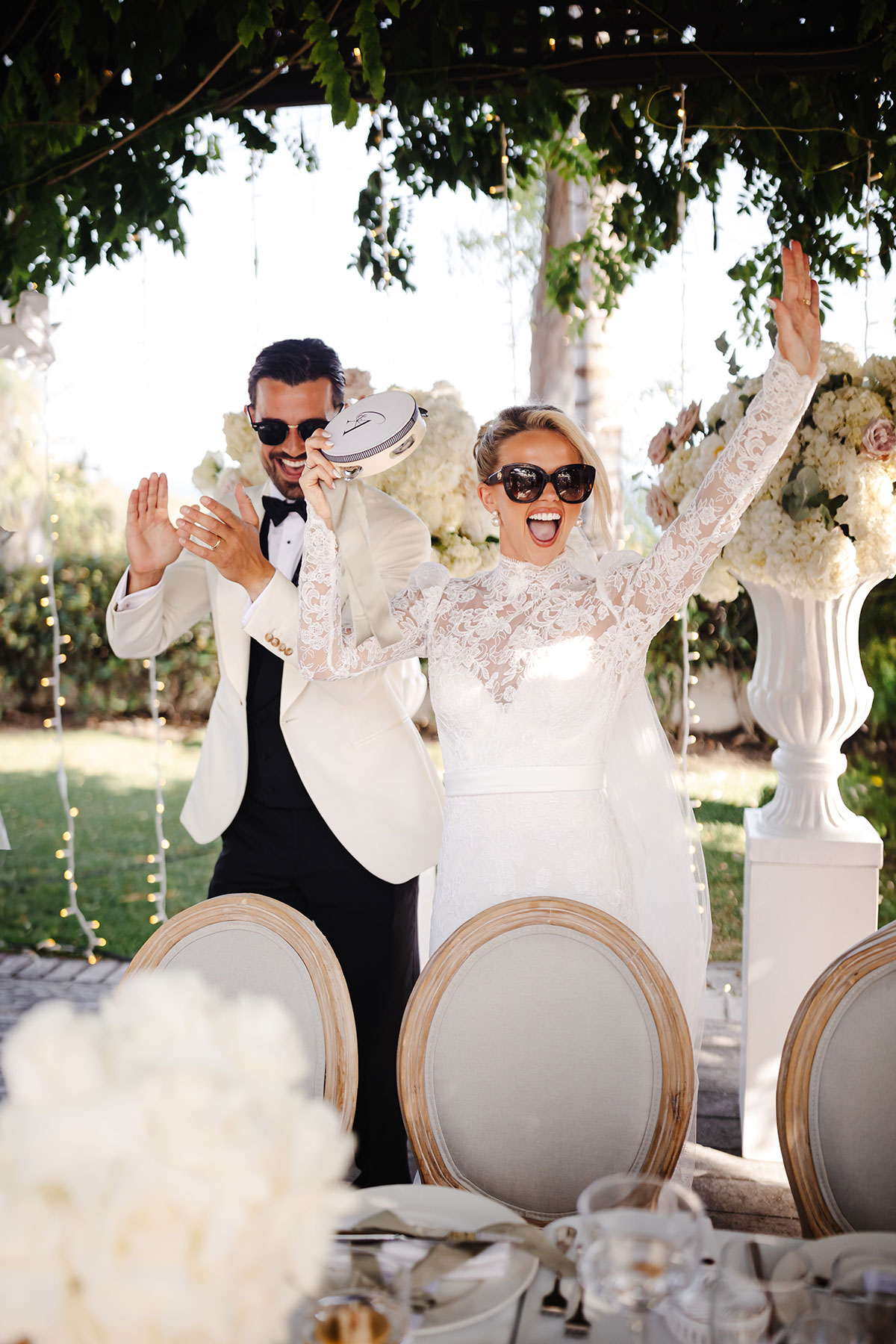 Bride and groom in sunglasses dance beside reception table under hanging foliage and fairy lights