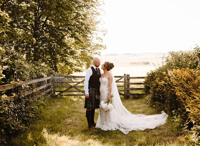 a bride and groom standing on grass with a wooden gate and fence behind them. There are trees on the left and a hedge on the right