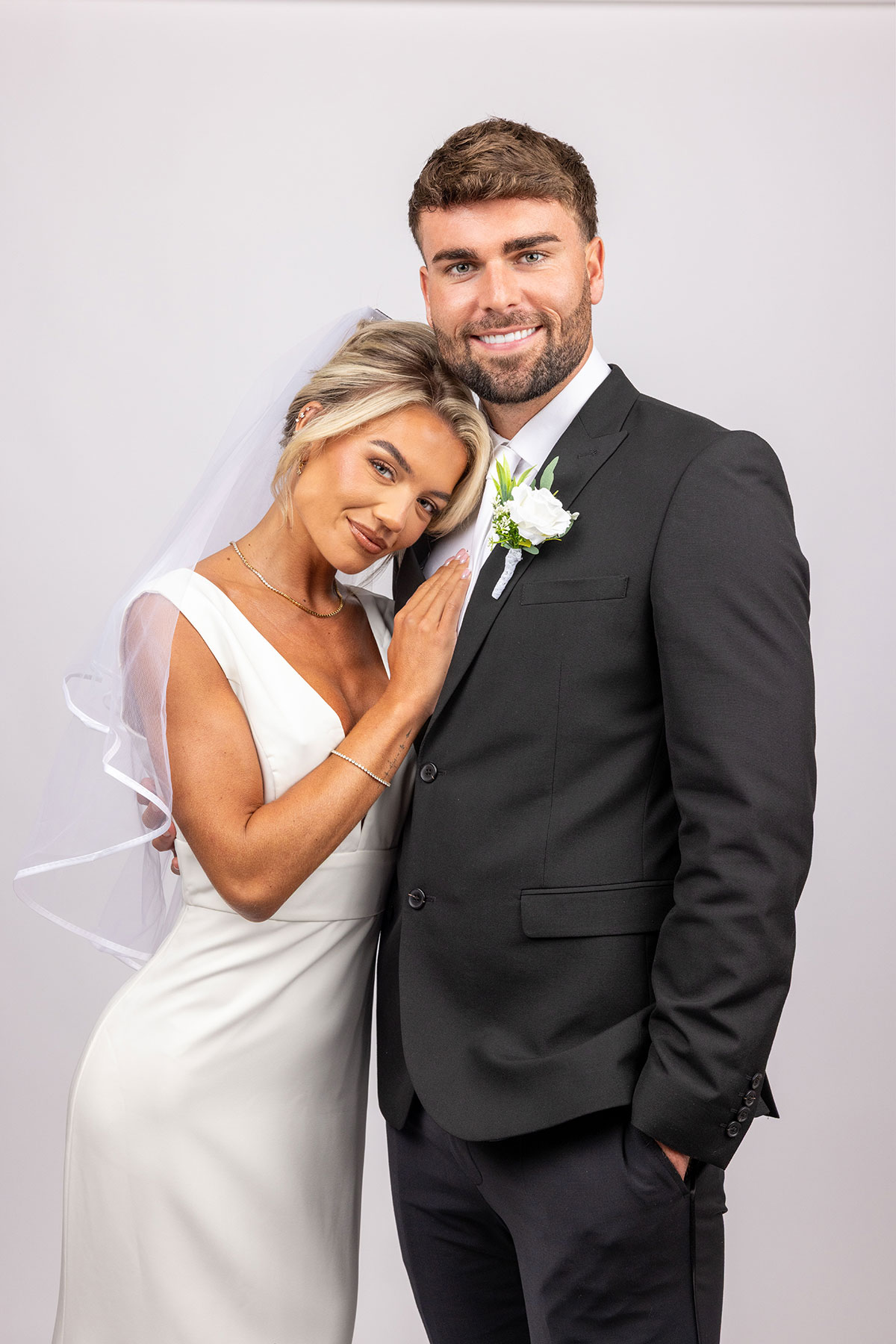 Bride in a simple satin wedding dress leans her head affectionately on the groom’s shoulder as they smile at the camera; he wears a black suit with white boutonnière against a plain studio backdrop