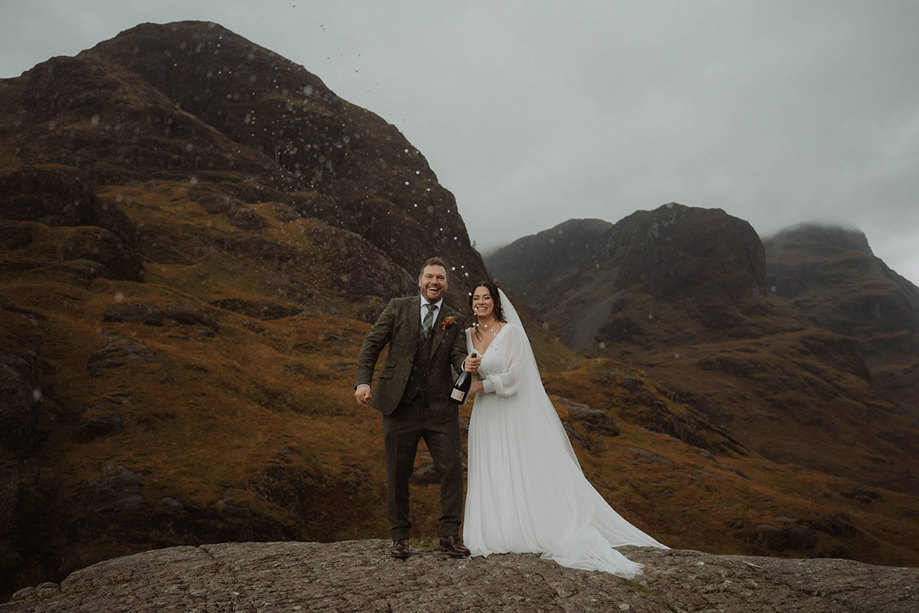 a bride and groom opening and spraying a bottle of champagne amid dramatic mountain scenery in Glencoe