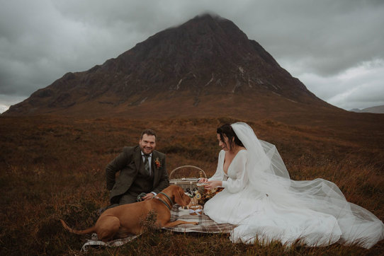 a bride and groom sitting on a tartan blanket with a dog while having a wedding picnic in the shadow of Buachaille Etive Mòr in Glencoe