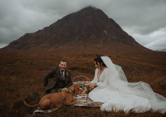 a bride and groom sitting on a tartan blanket with a dog while having a wedding picnic in the shadow of Buachaille Etive Mòr in Glencoe