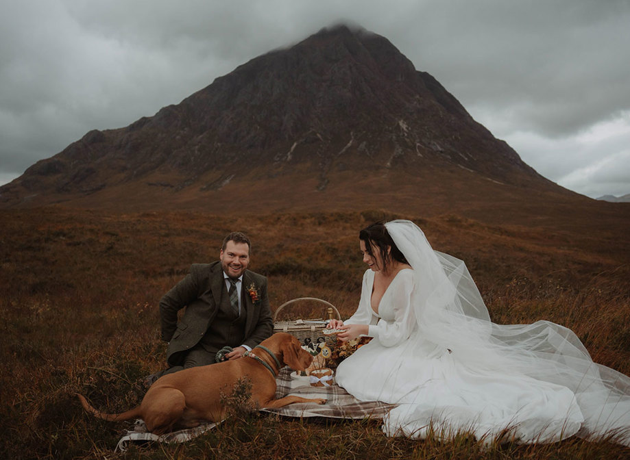 a bride and groom sitting on a tartan blanket with a dog while having a wedding picnic in the shadow of Buachaille Etive Mòr in Glencoe