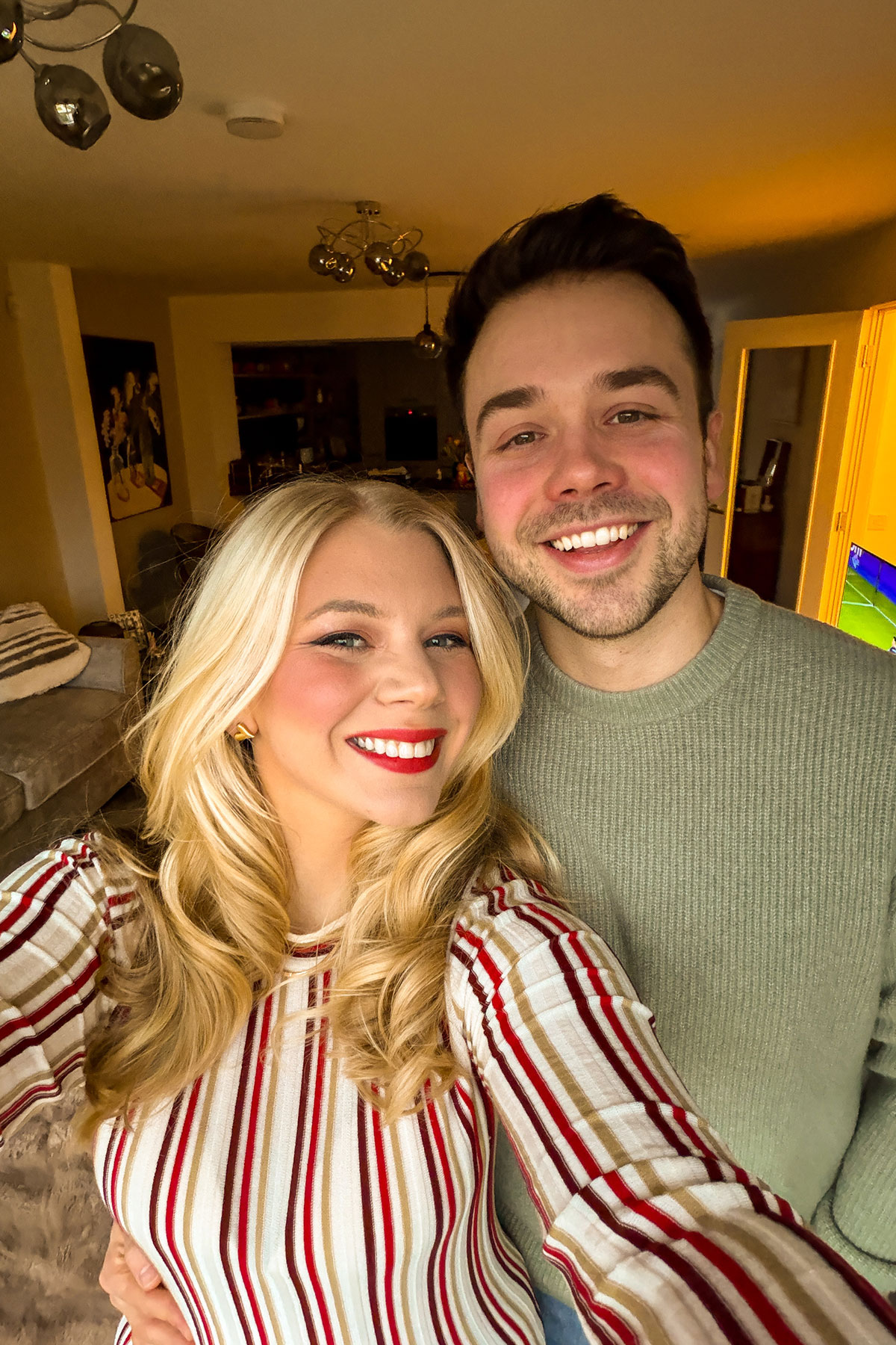Couple smiling selfie at home with warm lighting and cosy living room background