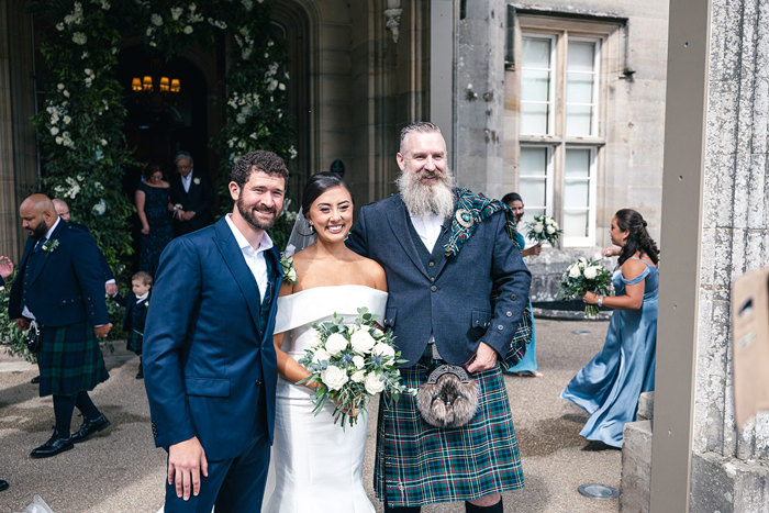 a smiling bride and groom pose outside Blairquhan Castle standing next to Andrew D Scott celebrant who is wearing a kilt while other guests walk in background