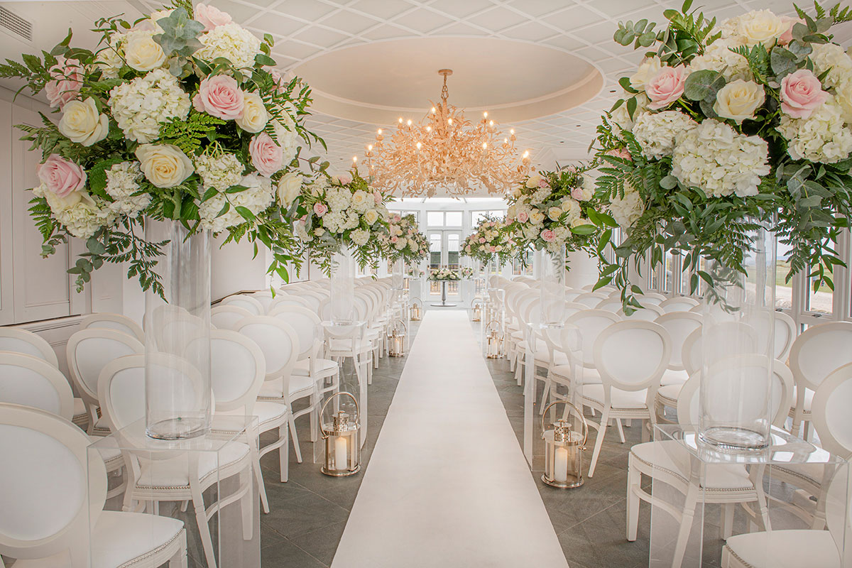 statement gold chandelier hangs from ceiling and there's a white runner in the middle surrounded by heightened pastel flowers and candle lanterns to create an aisle between two sides of white chairs 