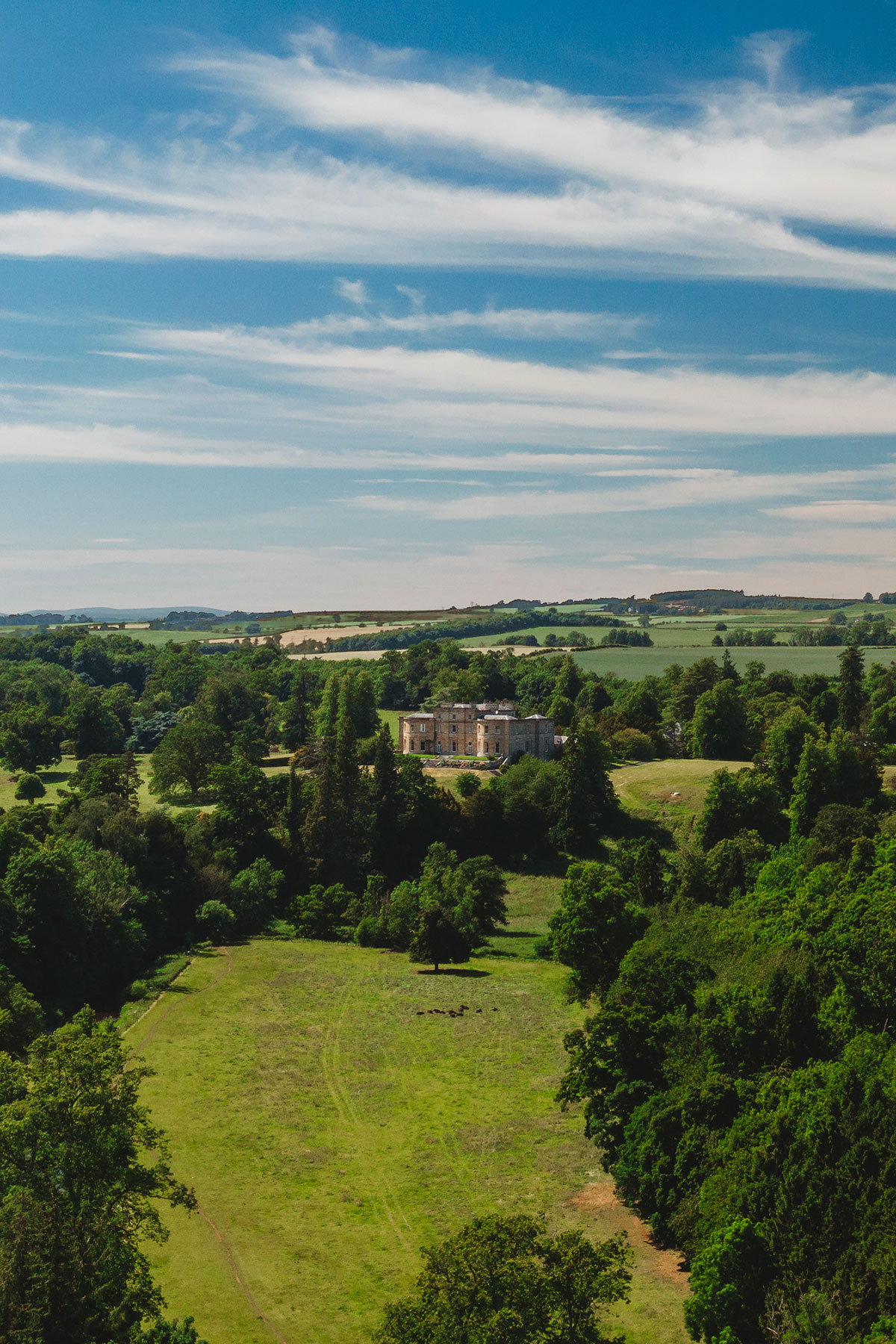 Aerial landscape showing Newton Don House set within rolling green fields, woodland and wide open rural scenery under a bright sky.