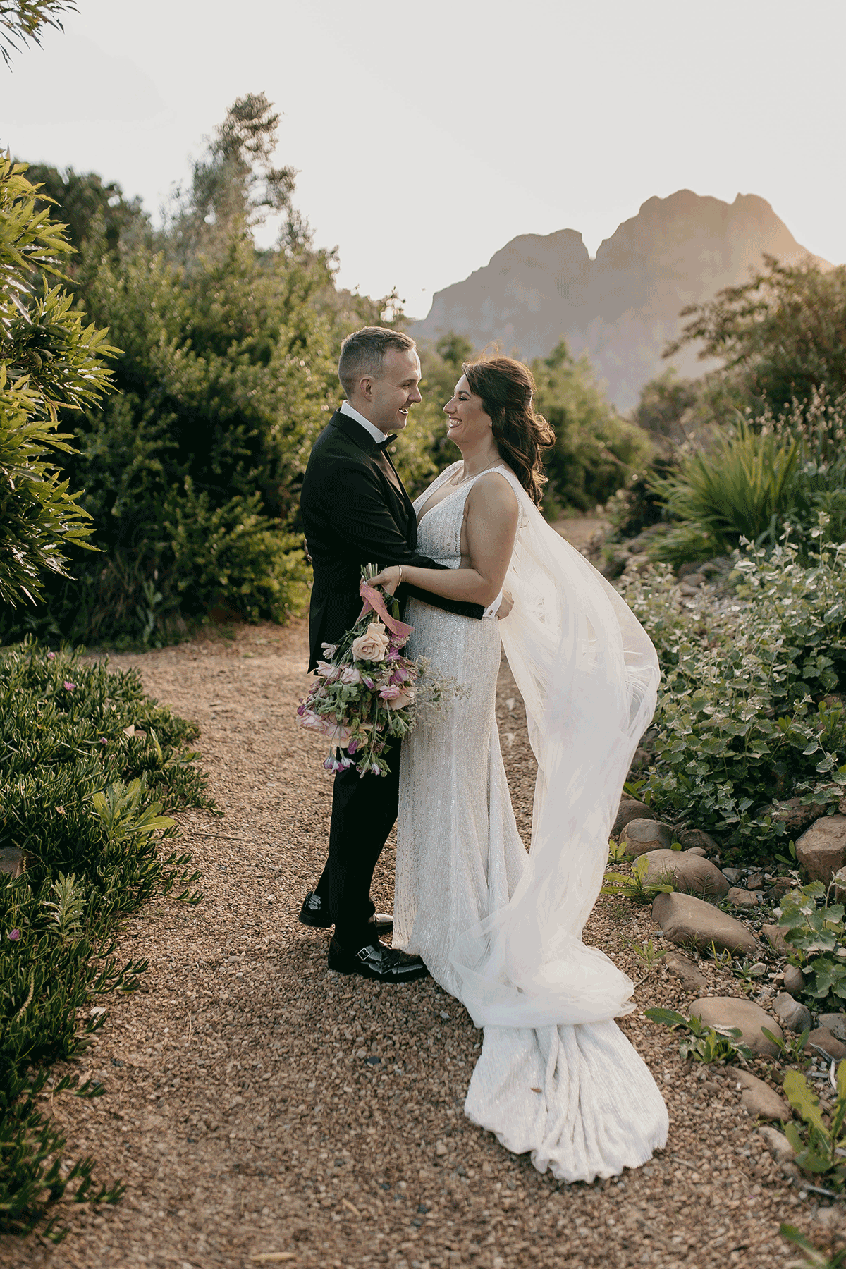 Bride and groom embracing in a garden at golden hour, with mountain scenery and soft greenery creating a romantic destination wedding backdrop