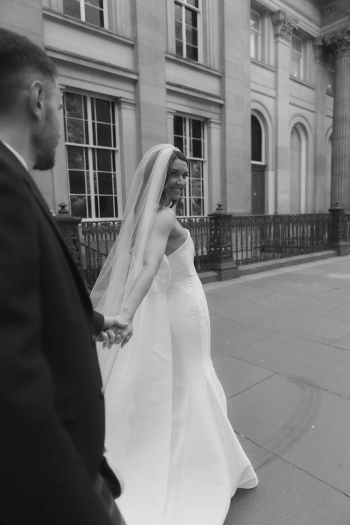 A bride looking over her shoulder at the camera as her hand stretches back to hold the groom's hand