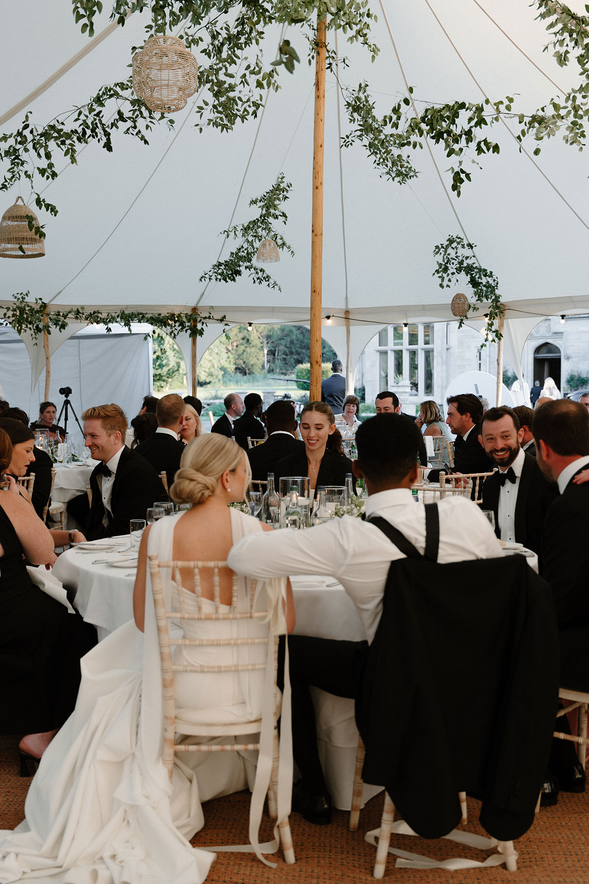 Wedding guests seated inside a sailcloth marquee reception with hanging greenery and woven lantern décor