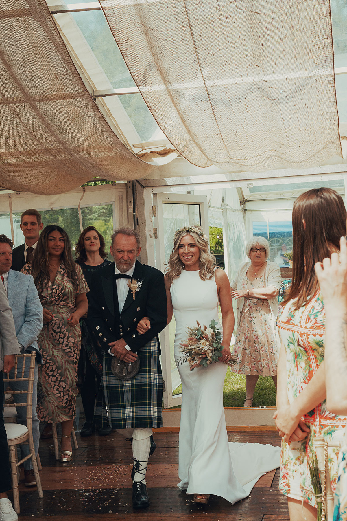 Bride walks down the aisle holding a bouquet, arm in arm with an older man in a kilt, inside a draped marquee as guests look on