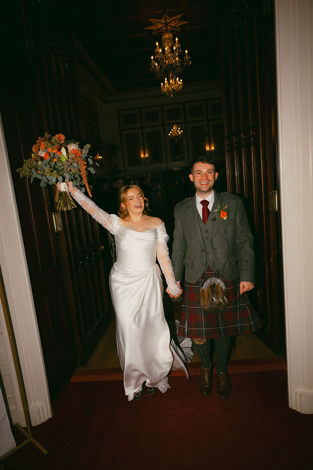 Newlywed couple entering reception with bouquet raised, groom in kilt, celebrating Scottish wedding day