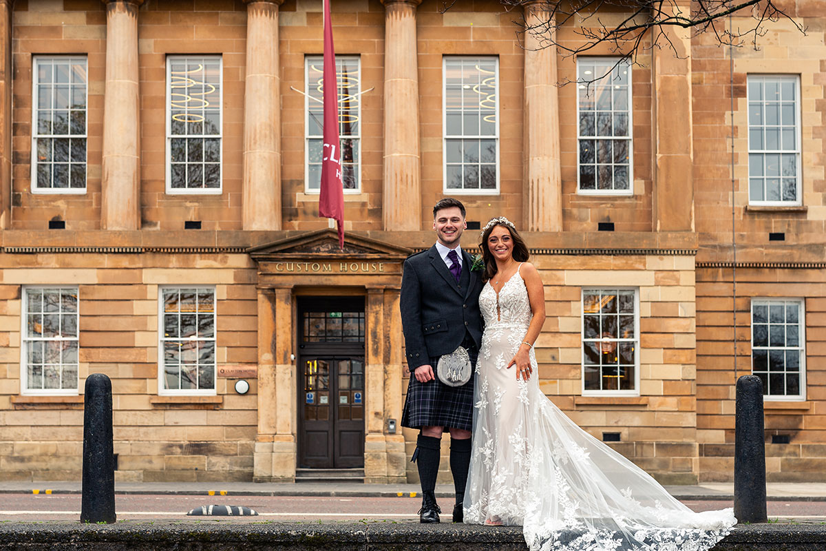 bride and groom stand in front of clayton hotel glasgow on wedding day wearing kilt and wedding dress