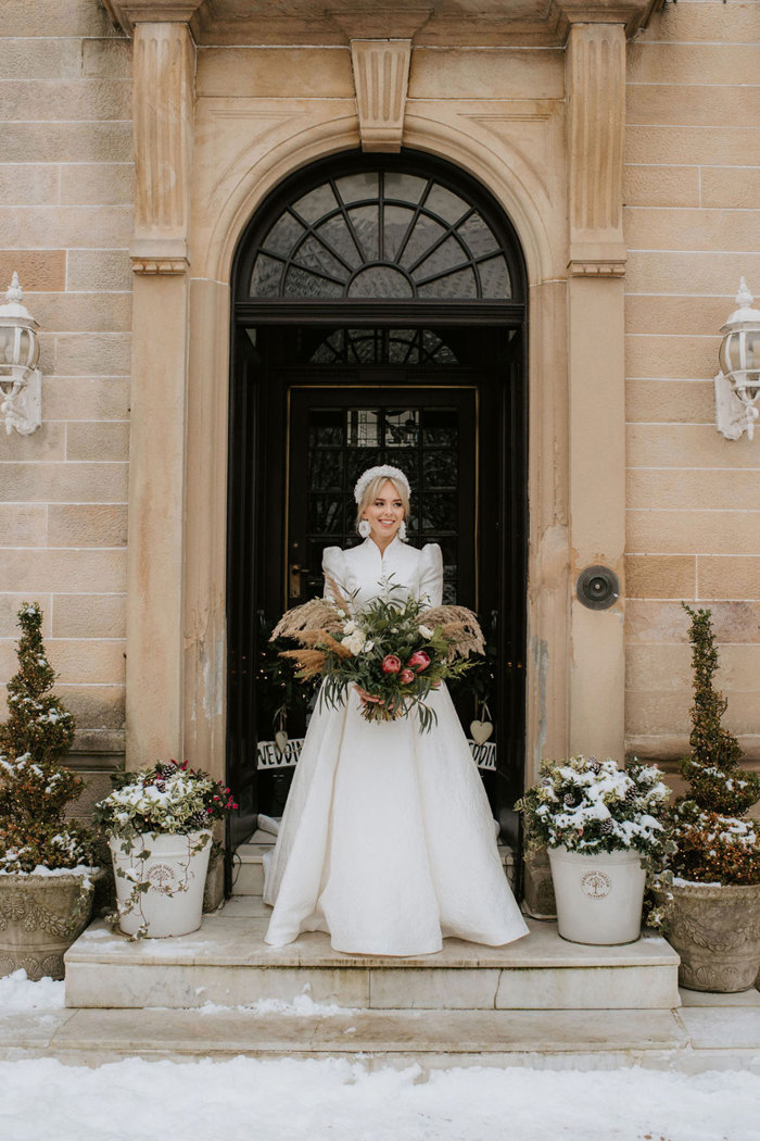 A bride wearing a textured dress with a matching high neck jacket standing on a doorstep holding a large bouquet