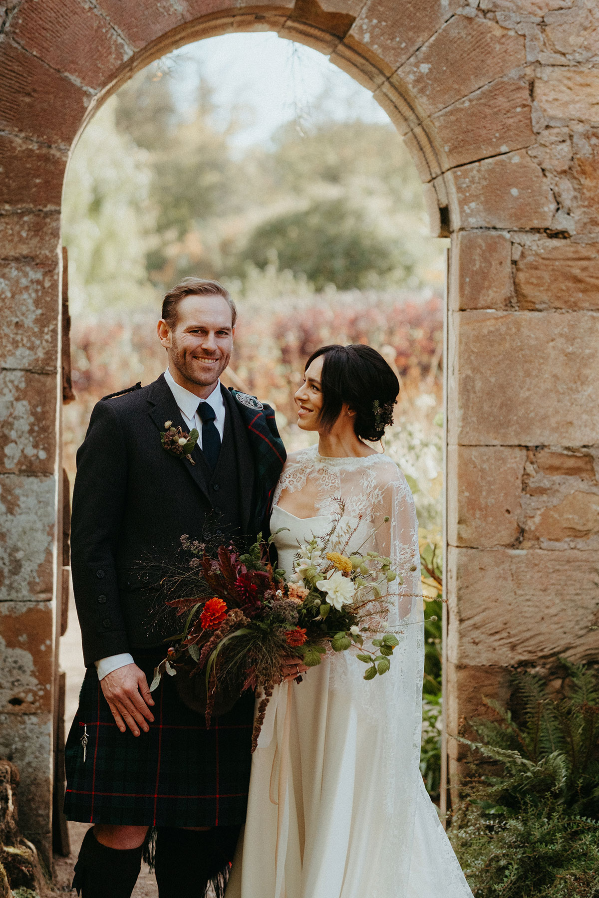 Bride and groom pose beneath a stone archway at Cambo Country House, holding an autumnal wedding bouquet.