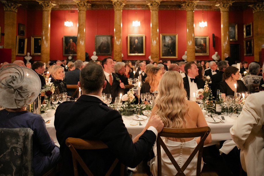 Room full of guests around tables - bride and groom with back facing camera looking at someone making a speech