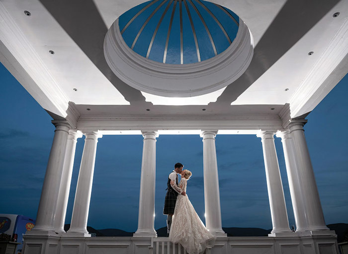 A bride and groom stand in between large white pillars with a blue night sky behind them