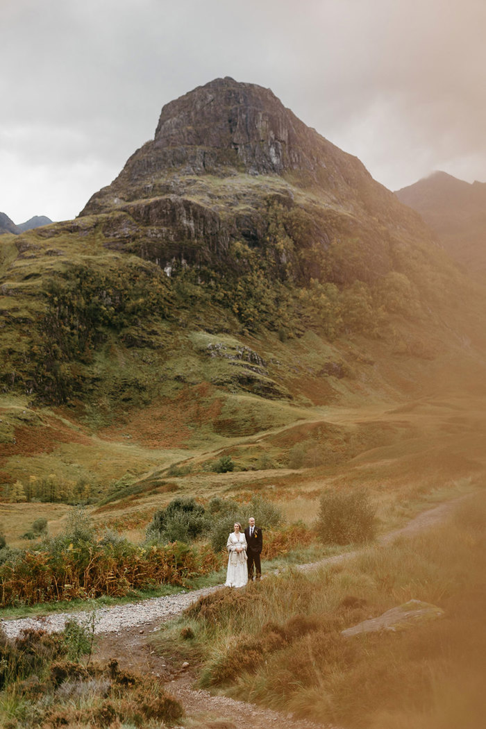 bride in white wedding dress and groom in black suit stand on small stone trail at the foot of tall hill