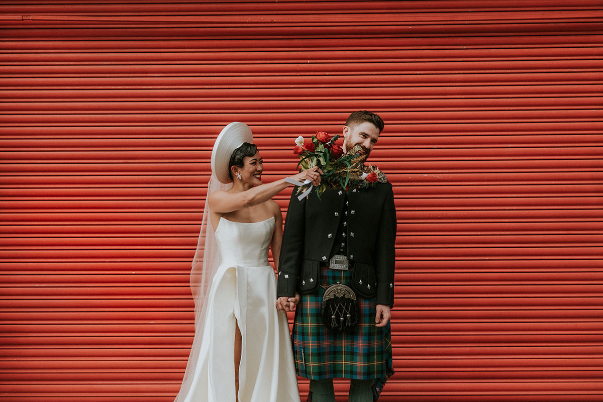 Bride playfully holding bouquet to groom’s face in front of red shutter at BAaD Glasgow wedding