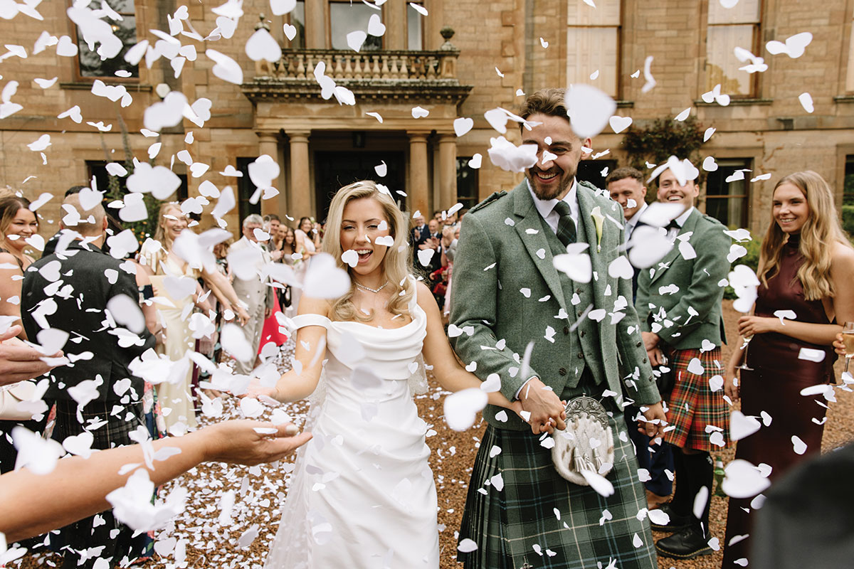 Newlyweds Lia and Louis smile as guests shower them with white confetti outside Cambo House in Fife.