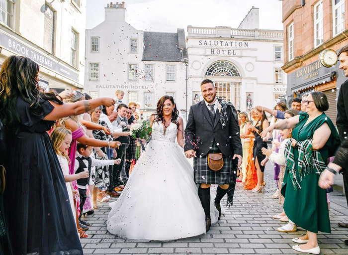 A bride in a ballgown wedding dress and a man in a kilt walking down a street as the wedding guests throw confetti over them