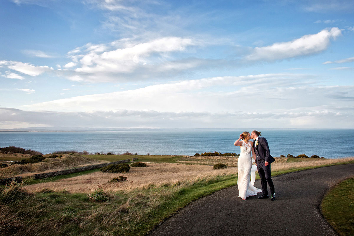 Newlyweds sharing a kiss on coastal road with sea views at Fairmont St Andrews