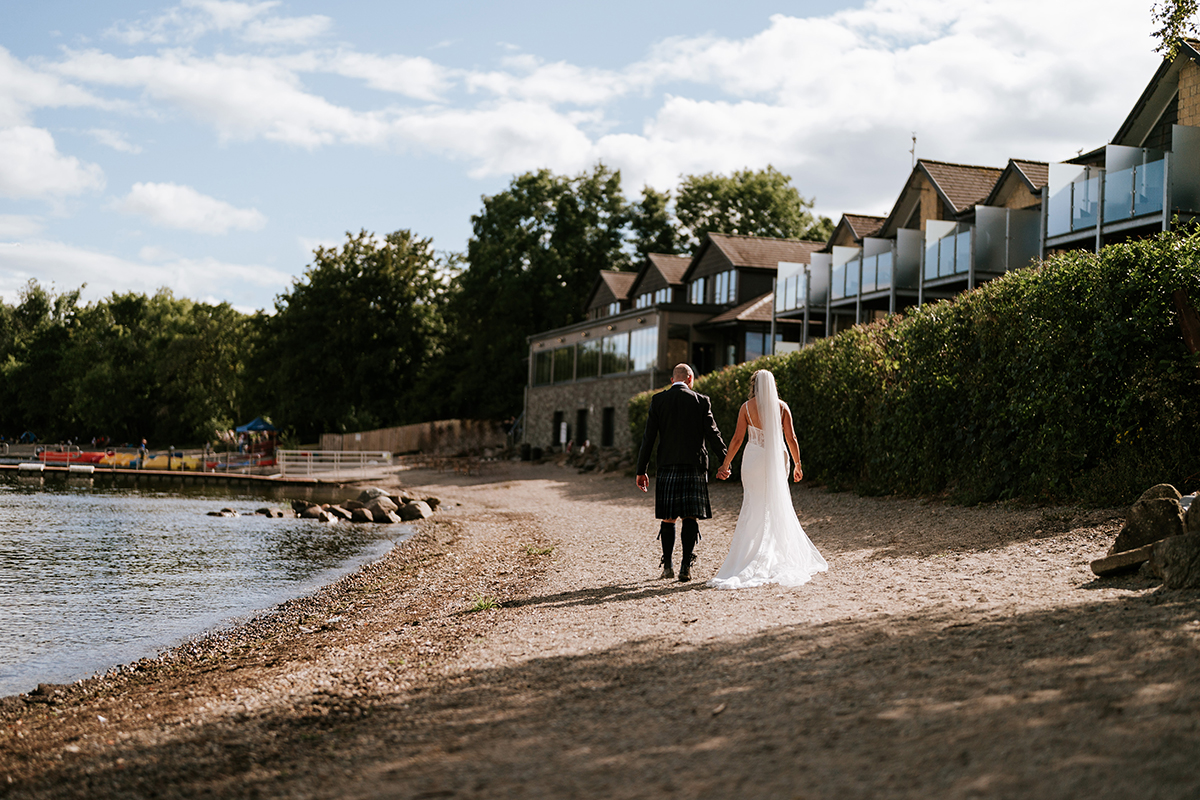 bride in trailing wedding dress and veil holds hands with groom in kilt outfit as they walk away from the camera along the sandy beach coast