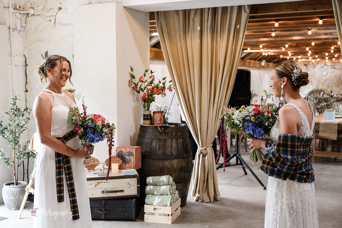 Two brides share a first look moment inside Blackshaw Barns, surrounded by floral arrangements and rustic décor with wooden barrels and draped curtains.