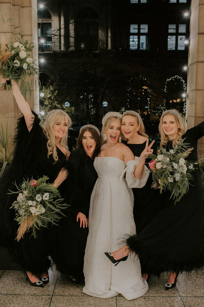 A bride wearing a sparkly white headband and a strapless dress with detached puff sleeves standing with bridesmaids wearing long black dresses and silver headbands