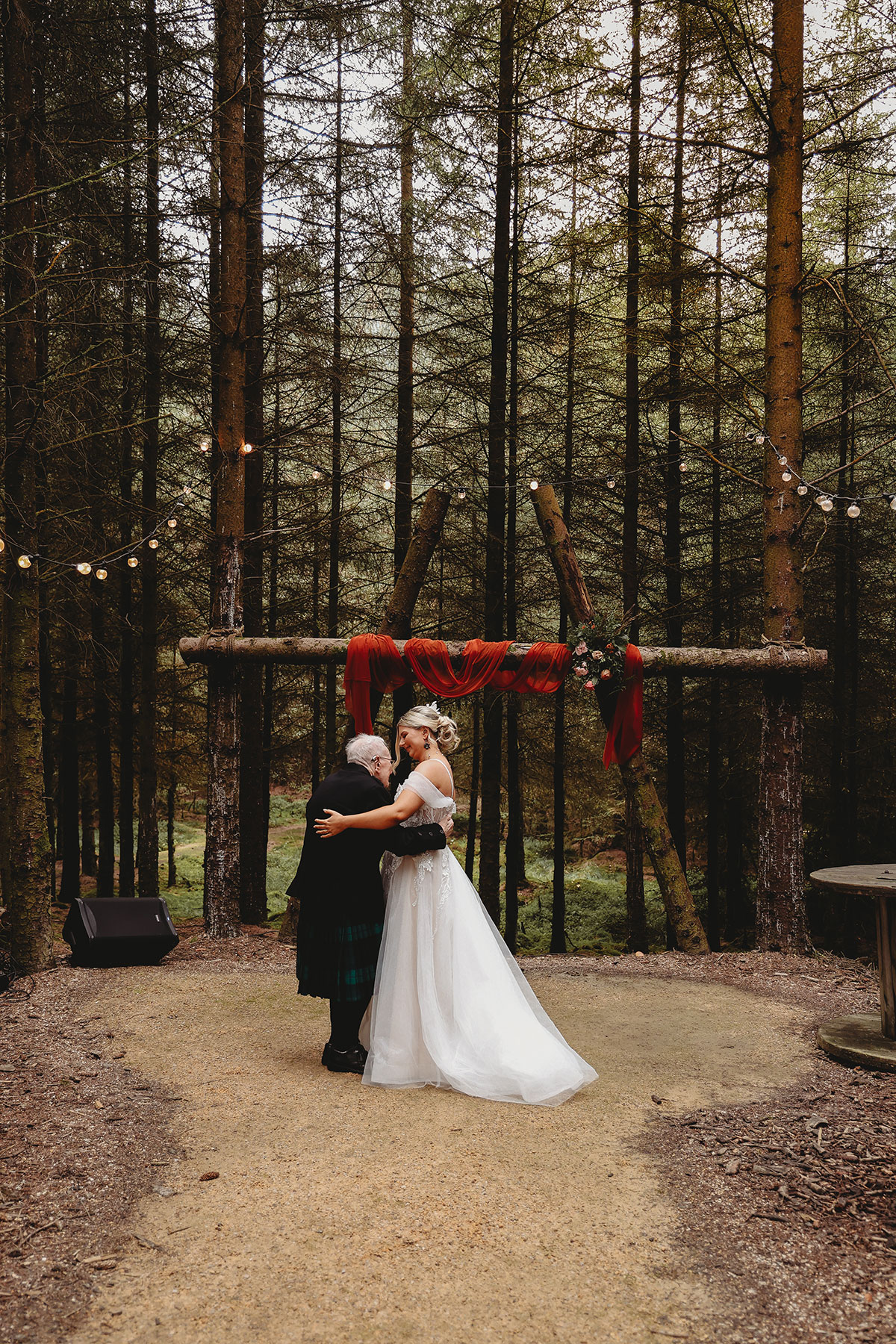 Bride sharing a hug with a family member beneath a rustic outdoor wedding arch during a Scottish woodland ceremony